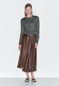 Hnědá plisovaná midi sukně v kombinaci s přízpusným šedým topem s dlouhým rukávem a měkkou texturou. Modelka má na sobě černé nazouvací sandály.