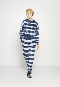 Pessoa a caminhar com um hoodie em tie-dye azul e branco e sweatpants combinando, usando sapatilhas verdes e pretas, olhando para o lado.