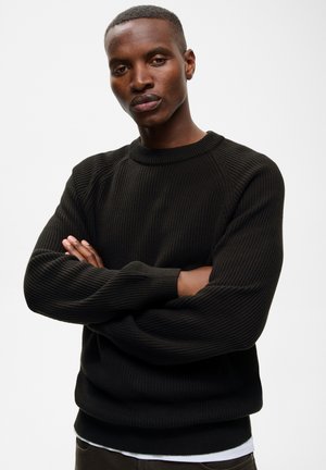 Homme aux cheveux courts portant un pull en maille noir et une chemise blanche en dessous, debout les bras croisés devant un fond uni.