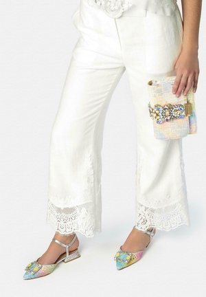 Mujer con pantalones blancos con encaje en los bordes y zapatos planos slingback multicolor con joyas, sosteniendo un pequeño bolso de mano pastel con joyas.