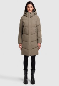 Olijfgroene pufferjas met capuchon, voorzien van een verticale quiltstructuur, twee zijzakken en drukknopen. Zwarte broeken en laarzen.