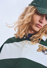 Grön och vit randig polotröja med krage och en broderad design som föreställer en karaktär. I sällskap av en grön keps med en logotyp.