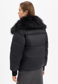 Kobieta z krótkimi włosami, ubrana w czarną puchową kurtkę z dużym czarnym futrzanym kołnierzem, w połączeniu z szarym spódnicą, stoi bokiem na białym tle.