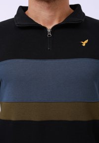 Černý top s čtvrtinovým zipem s horizontálními pruhy v modré a olivové barvě, s malým zlatým logem ptáka na levém prsu. Měkká textura.