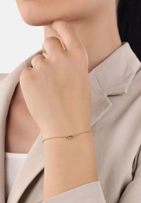 Brazalete delicado de cadena de oro con un pequeño grupo de piedras oscuras y claras, usado en una muñeca contra ropa beige.