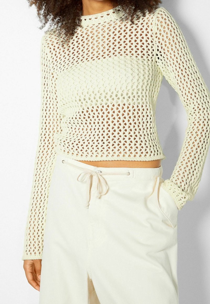 Stickad långärmad topp i ljuskräm, med ett diamantsmönster, ribbad halsringning och åtsittande ärmslut. Kombinerad med krämfärgade byxor med snörning.
