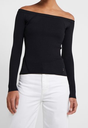 Kobieta ubrana w dopasowaną czarną prążkowaną bluzkę z długim rękawem i odkrytymi ramionami, zestawioną z białymi spodniami z wysokim stanem, na jednolitym tle.