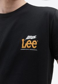 Inimene, kes kannab musta T-särki oranži "Lee" logoga ning valget lippu, millel on kiri "1889", ning rindkere peal tekst "American Original".