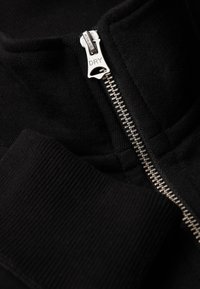 Sort zip-jacke med sølv lynlås og ribbet krave. Blødt stof med minimalisk design og fokus på detaljerne.
