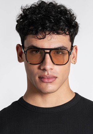 Mladić s kovrčavom crnom kosom nosi velike, kvadratne sunčane naočale smeđe nijanse i crnu rebrastu majicu na jednostavnoj pozadini.