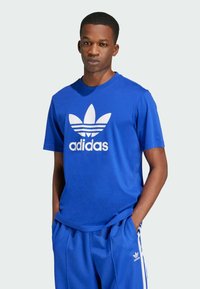 Blue cotton t-shirt featuring a white Adidas logo and trefoil graphic on the front. Short sleeves and round neckline. Simple, sporty design.