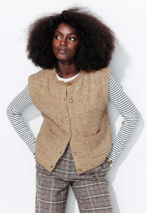 Femme noire avec coiffure afro naturelle portant un gilet beige boutonné, un haut à manches longues rayé et un pantalon à carreaux, se tenant les mains sur les hanches.