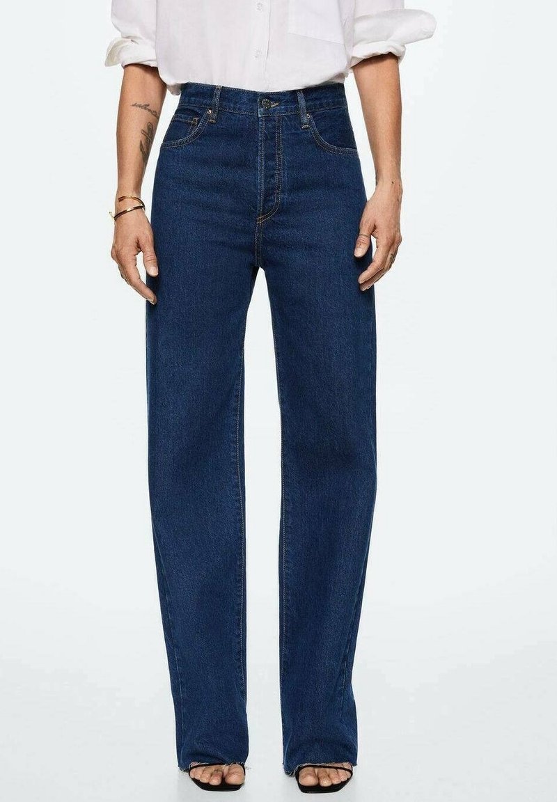 Personne portant un jean droit taille haute bleu foncé, une chemise blanche aux manches retroussées et des sandales noires à bouts ouverts, debout devant un fond blanc.