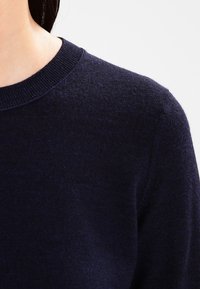 Mörkblått stickat tröja med rund halsringning. Mjuk textur och subtil lyster. Har ribbade ärmslut och fåll, vilket framhäver en figursydd design.