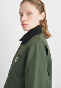 Groene jas met een zwarte, gestructureerde kraag. Bevat een zijzak, gladde stof en een patch-logo op de linkertepel.