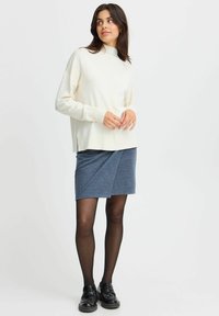Krämfärgad stickad tröja med hög halsringning, kombinerad med en figursydd blå omlottskjol. Svarta skor fulländar outfiten. Minimalistisk design.
