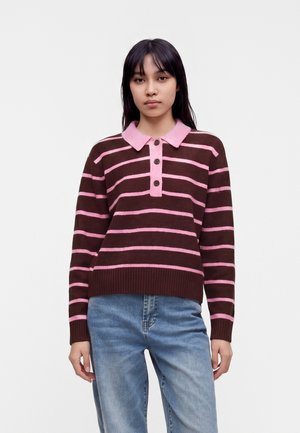 Jeune femme portant un pull rayé brun et rose à manches longues avec un col boutonné et un jean bleu clair, debout sur fond blanc.