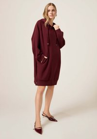 Burgundowa, oversized sukienka z kapturem, z przednią kieszenią, regulowanym sznurkiem oraz zatrzaskami na końcu; miękka tkanina o gładkiej fakturze.