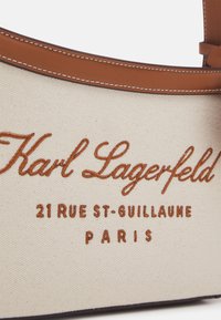 Gros plan d’un sac en tissu beige avec garniture en cuir marron, texte brodé « Karl Lagerfeld 21 Rue St-Guillaume Paris » en fil marron.