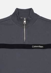 Szara bluza na zamek z wysokim kołnierzem, rypsowym wzorem i czarnym paskiem z białym logo "Calvin Klein". Zamek błyskawiczny z srebrnym wykończeniem.