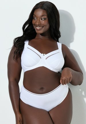 Lächelnde Frau mit langen Haaren, die einen weißen BH und passende hoch taillierte Unterwäsche trägt und am hellen Hintergrund den Bund justiert.