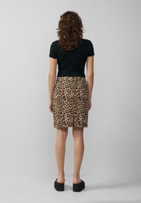 Frau mit lockigem Haar, die ein schwarzes kurzärmliges Oberteil, einen Rock mit Leopardenmuster und schwarze Plateau-Sandalen trägt, steht vor einem einfarbigen grauen Hintergrund.
