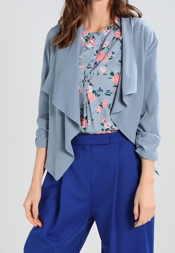 Ljusblå draperad jacka över en blommig blus, parat med lösa, livfullt blå byxor. Slät tyg med lagerdesigndetaljer.