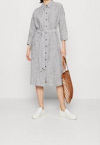 Gestreiftes Hemdblusenkleid mit einem Gürtel in Grau und Weiß, mit Kragen und Drei-Viertel-Ärmeln. Kombiniert mit einer geflochtenen beige Handtasche und weißen Sneakers.