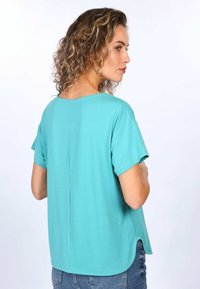 Tuerkisfarbenes Kurzarmoberteil mit rundem Ausschnitt, lockerer Passform und Rückennähtedetail, kombiniert mit blauer Jeans. Glatte Stofftextur.