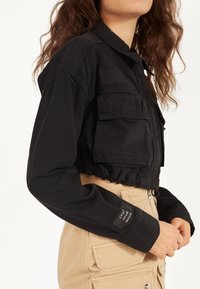 Krátká černá bunda z bavlny, s dvěma našitými kapsami, zapínáním na knoflíky a elastickým lemem. Na rukávu je štítek.