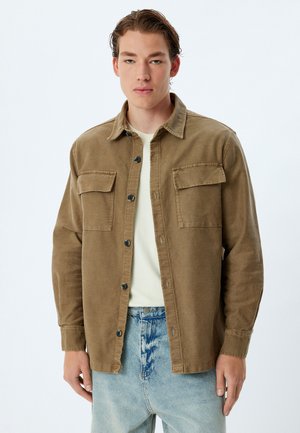 Bruine corduroy overhemdjas met twee borstzakken, knoopsluiting en een relaxte pasvorm, gecombineerd met een lichtgekleurde T-shirt en denim jeans.