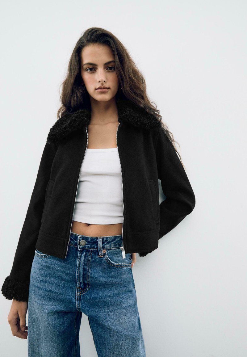 Giacca nera con collo peloso, chiusura con zip e tasche laterali, abbinata a un top bianco senza maniche e jeans in denim blu a vita alta.