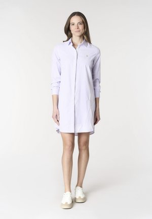 Giovane donna in piedi che indossa un vestito camicia a righe lilla chiaro e scarpe da ginnastica bianche su sfondo bianco.