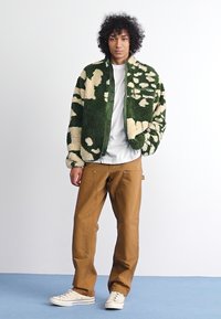 Jaqueta bomber felpuda, verde e com padrão creme, com gola alta, combinada com calças castanhas e ténis brancos. O modelo está em cima de uma superfície azul.