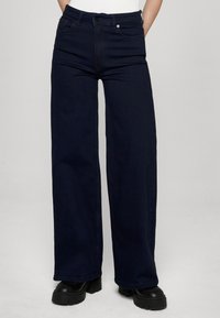 Tmavomodré džínsové nohavice s širokými nohavicami, vyrobené z denimu, s predným zapínaním na gombíky, piatimi vreckami a jemnými švami.