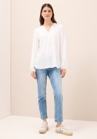 Hvid langærmet bluse med V-hals og knaplukning, parret med lyseblå lige ben jeans og beige sneakers med blå accenter.
