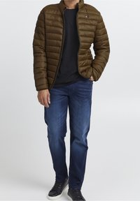 Brun dunjakke med lynlås foran, quiltet design og høj krave, kombineret med en sort skjorte og blå denimjeans. Sorte sneakers fuldender looket.