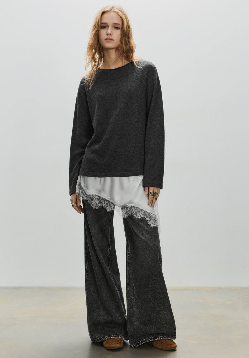 Oversized mørkegrå sweater lagdelt over en hvid satin top med blonderkant, kombineret med vidde ben falmede sorte jeans og brune sko.