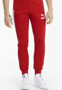 Red athletic pants with white side stripes, made of a smooth material, featuring a fitted design and a Puma logo on the front.