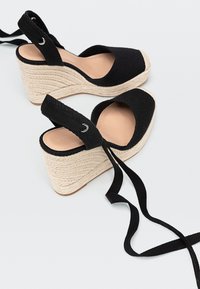 Espadrilles à plateforme en tissu noir avec des semelles en corde tressée beige et de longues lanières noires autour de la cheville sur fond blanc.