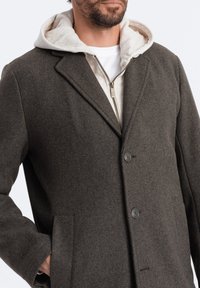 Manteau en laine gris foncé avec un col à revers, deux boutons et poches latérales, superposé sur un sweat à capuche crème zippé. Texture douce, coupe ajustée.
