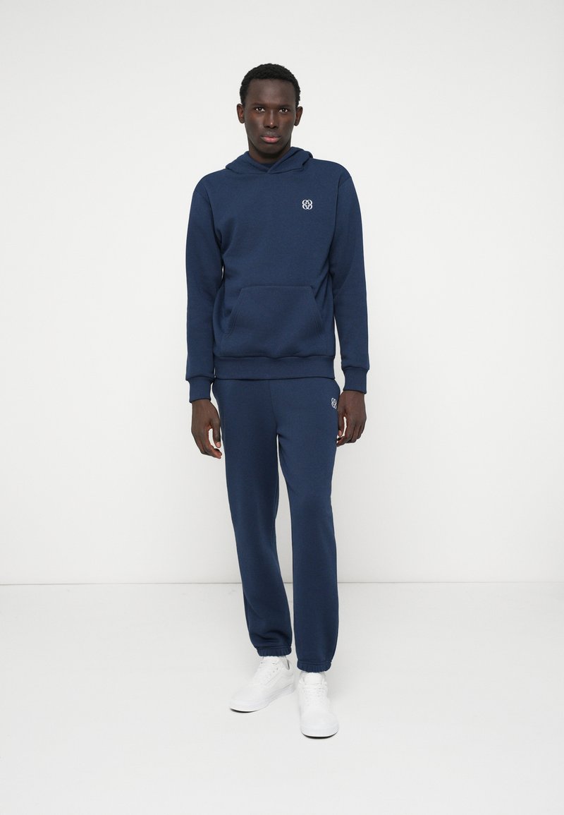 Navy blue hoodie and sweatpants set made of soft fabric, featuring a front pocket, ribbed cuffs, and an embroidered logo on the chest.