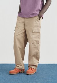 Άτομο που φοράει μπεζ παντελόνι τύπου cargo, πορτοκαλί δερμάτινα παπούτσια με κορδόνια και μοβ πουκάμισο, με το ένα χέρι στην τσέπη, πάνω σε μπλε χαλί.