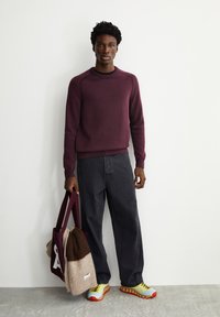 Bordeauxfarbener Strickpullover mit geripptem Ausschnitt, dunkelgraue Jeans, mehrfarbige Sneakers und eine Tasche mit einer braunen und hellbraunen Decke.