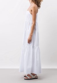 Bílá maxi šaty s tierováním a ramínky typu spaghetti, vyrobené z hladkého materiálu. Kombinované se světle šedými sandály s minimalistickým designem a přezkami.