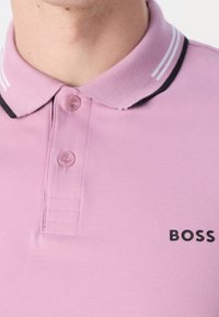 Gros plan sur une personne portant un polo rose avec un col rayé noir et blanc et un logo "BOSS" sur la poitrine.