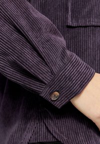 Paarse corduroy overhemdmanchet met verticale ribbels, een knoopsluiting en een relaxte fit. Details omvatten opvallende stiksels.
