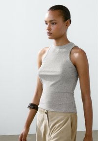 Grijze geribde tanktop met een hoge halslijn en mouwloos design, gecombineerd met lichtgekleurde broek. Opvallende accessoire: donkere armband om de arm.