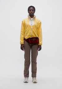 Άτομο στέκεται προς τα εμπρός φορώντας ένα κίτρινο κοντό μπουφάν με φερμουάρ, καφέ παντελόνια, λευκά παπούτσια και κόκκινη ζώνη μέσης.