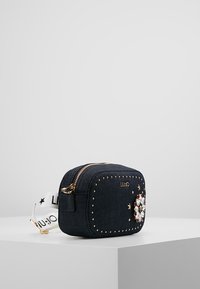 Bag in denim con borchie dorate, decorazioni floreali e una tracolla bianca. Forma rettangolare, chiusura con zip, design compatto.